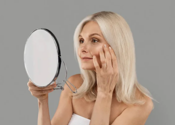 Skin Aging. Attractive Mature Woman Holding Mirror And Looking At Her Wrinkles Near Eyes, Beautiful Senior Female Checking Face, Noticing Age Changes, Standing Wrapped In Towel On Grey Background